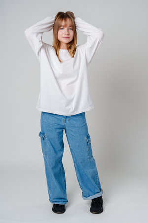Young woman in a casual white long-sleeve shirt and loose denim pants poses against a light gray backgroundの写真素材