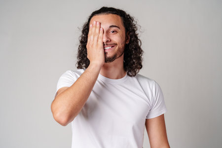 A young man playfully covering one eye with his hand while smiling in a neutral-toned indoor settingの写真素材
