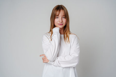Young woman in a white long-sleeve shirt thoughtfully poses with a hand on her chin in a minimalistic settingの写真素材