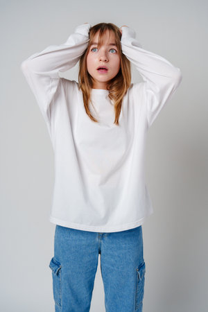 A teen girl in a loose white sweater looks surprised with her hands on her head against a plain gray backdropの写真素材