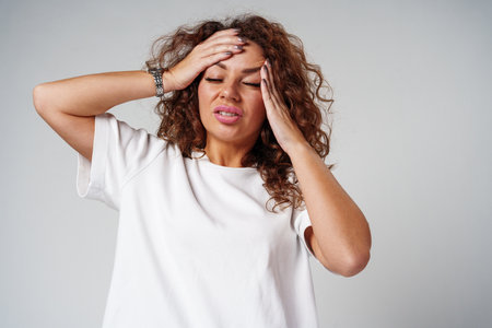 Woman with curly hair holds her head in discomfort, expressing concern or stress in a neutral backdropの写真素材