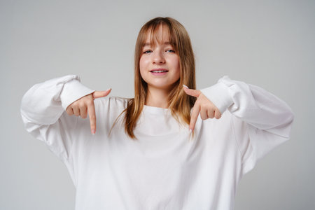 A young girl in a white long-sleeve shirt smiles while gesturing downwards in a light-colored roomの写真素材