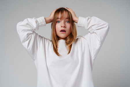 A teen girl in a loose white sweater looks surprised with her hands on her head against a plain gray backdropの写真素材