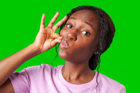 A young woman in a pink shirt makes a shushing gesture against a bright green background
