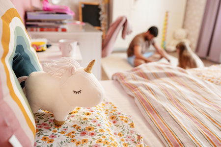 A white unicorn plush toy sits on a bed with a rainbow pillow in a bedroom, blurred figures in the backgroundの写真素材