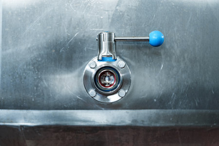 A stainless steel valve with a blue handle is attached to a large container