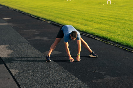 Man stretching on a running track in the early morning light as grass glistens nearbyの写真素材