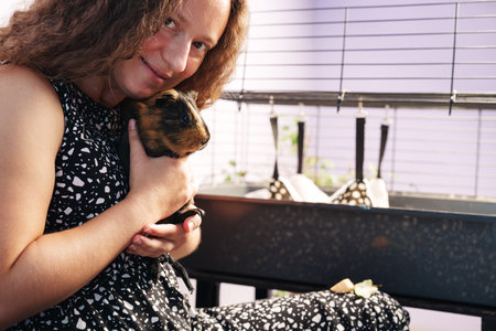 Young girl holds a guinea pig in her arms while sitting next to a cage in a cozy indoor settingの写真素材
