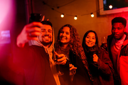 Group of friends enjoying a night out at a cozy bar, capturing memories with cheerful selfies while holding drinksの写真素材