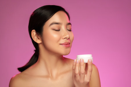 Woman enjoying skincare product with serene expression under soft pink lightの写真素材