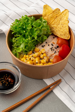 Healthy bowl with grilled chicken, lettuce, corn, cherry tomatoes, tortilla chips, and sauce on a tableの写真素材