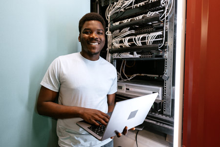 Young professional managing servers with laptop while smiling in a data center during daytime