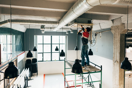 Construction worker performing maintenance on ceiling equipment in modern workspace during daylight hoursの写真素材