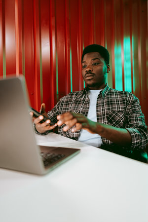 Focused young man working on a laptop while using a smartphone in a vibrant workspaceの写真素材