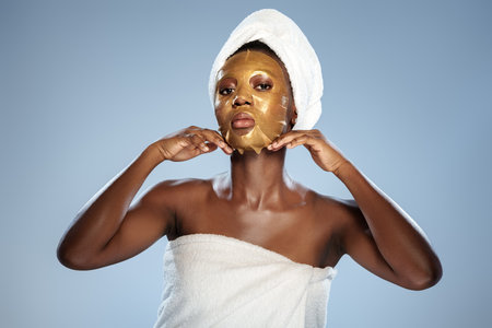 Woman applies gold facial mask while wrapped in a towel in a serene indoor settingの写真素材