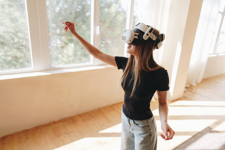 Young woman experiencing virtual reality in a sunlit room with large windows and wooden flooring