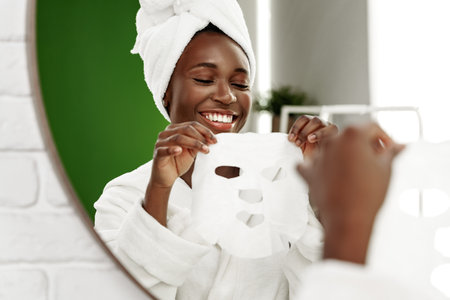 Woman applying a facial mask while smiling in a bathroom setting with a mirror reflectionの写真素材