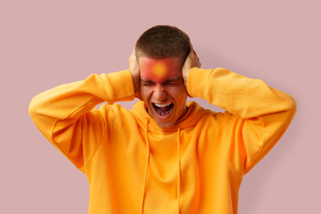 Young man expressing frustration with hands on head in a casual orange hoodie against a pink backgroundの写真素材