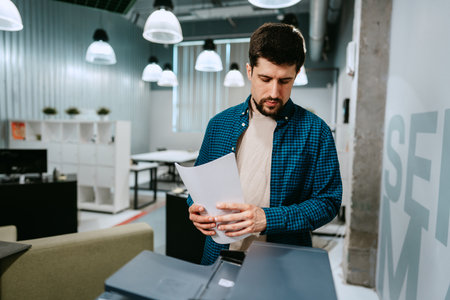 Man using a printer in a modern office space during the day while reviewing printed documents