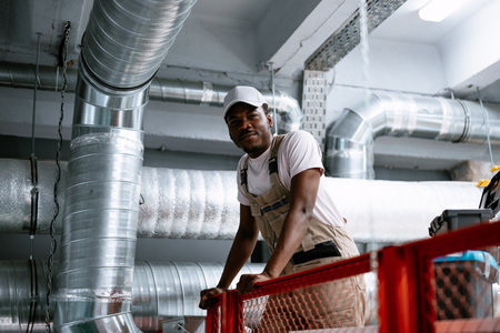 Worker on elevated platform inspecting ventilation system in industrial facility during daytimeの写真素材