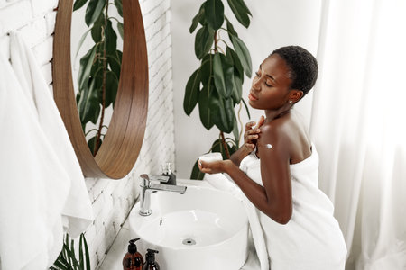 Woman applying moisturizer in a serene bathroom setting with plants and natural lightの写真素材