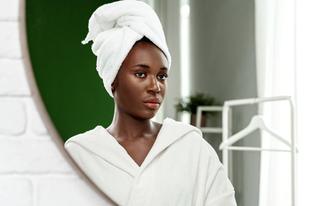 Woman in a white robe and towel gazes in the mirror after a refreshing shower in a cozy bathroomの写真素材