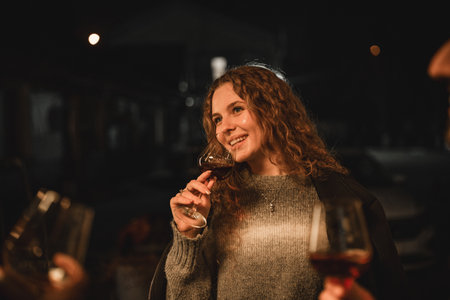 Woman smiling while holding a glass of wine at a nighttime gathering with friends outdoorsの写真素材