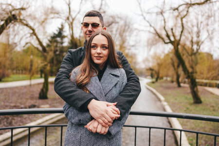 Couple embraces on a scenic bridge during a cloudy day in a tranquil park with trees in early springの写真素材
