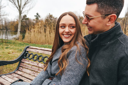 Couple enjoying each others company on a park bench during a chilly autumn afternoon by the waterの写真素材