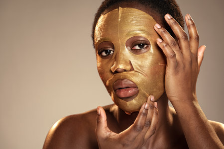 Woman applying a golden facial mask in a studio setting with soft lighting during a self-care routineの写真素材