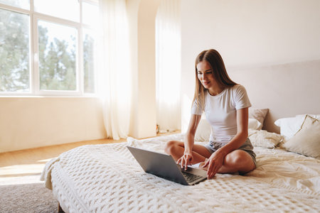 Young woman working on laptop while sitting on bed in bright, cozy bedroom during daytimeの写真素材