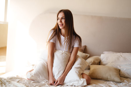 Smiling woman sitting on bed surrounded by pillows in a cozy, sunlit bedroom during the morningの写真素材