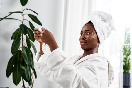 Woman in a towel applying skincare treatment at home in a bright, modern bathroom with indoor plantsの写真素材