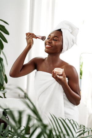 Woman applying skincare routine in bright bathroom, wrapped in towel with fresh plants aroundの写真素材