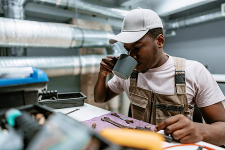 Man enjoying a warm drink while working on tools in a modern workshop setting during the dayの写真素材