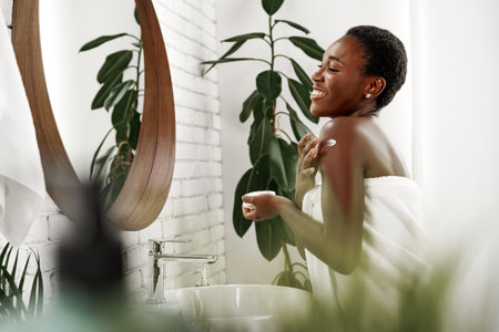 Woman applying lotion and enjoying self-care in a bright bathroom with plants and a large mirrorの写真素材