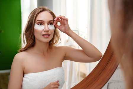 Woman applying under-eye patches in a bright, relaxing bathroom setting during daytimeの写真素材