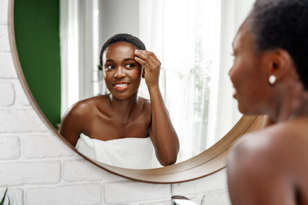 Woman applying skincare product in a well-lit bathroom while looking in a round mirrorの写真素材