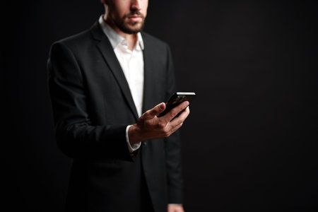 Businessman dressed in suit checking smartphone while standing against a dark backgroundの写真素材
