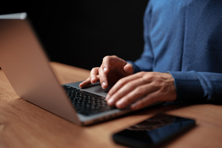 Hands typing on a laptop while a smartphone rests on a wooden desk during evening hoursの写真素材