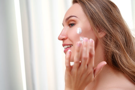 Woman applying skincare cream in a bright room near a window during daytime while smilingの写真素材