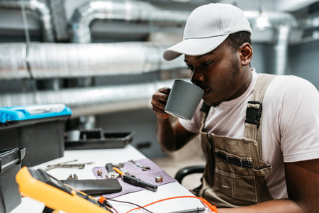 Young man enjoying a coffee break while working on electrical equipment in a modern workshop settingの写真素材