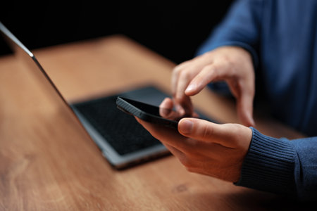 Person using smartphone while sitting at wooden desk with laptop during evening hours in a cozy settingの写真素材