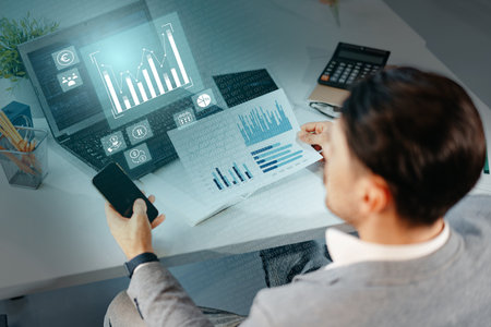 Individual analyzes financial data on laptop while reviewing printed reports and using smartphone in modern office settingの写真素材