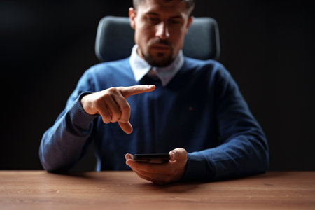 Man interacting with smartphone at a desk during a low-light setting in a modern workspaceの写真素材