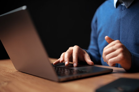Focused individual working on a laptop at a wooden desk in a dimly lit room during evening hours