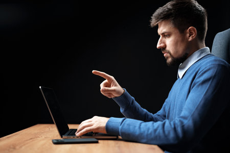 Focused man working on laptop in a dimly lit room, engaged in a serious task while seated at a wooden deskの写真素材