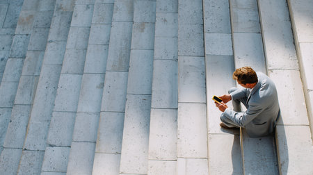 Businessman sitting alone on stairs checking smartphone in urban setting during daytimeの素材