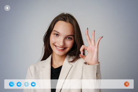 Young woman in a light suit making an okay gesture in a professional setting against a neutral backgroundの写真素材