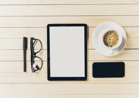 Workspace setup with tablet, glasses, pen, phone, and coffee on wooden tableの写真素材